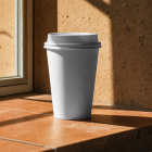 A blank white paper coffee cup with a lid standing on a terracotta tiled windowsill in warm, direct sunlight.
