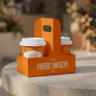 An orange cardboard drink carrier holding two paper coffee cups with white lids, sitting on a round concrete table in an outdoor café setting.