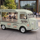 Front-three-quarter view of a vintage light green coffee and tea food truck mockup with an open hatch and staff working inside, parked in an urban courtyard with outdoor seating.