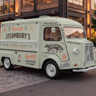 Side-angle view of a vintage light green food truck mockup parked along a cobbled street curb in front of modern brick buildings at dusk.