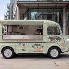 Side profile of a vintage light green coffee food truck mockup with open side and rear hatches, parked on a wide paved sidewalk near modern glass office buildings.