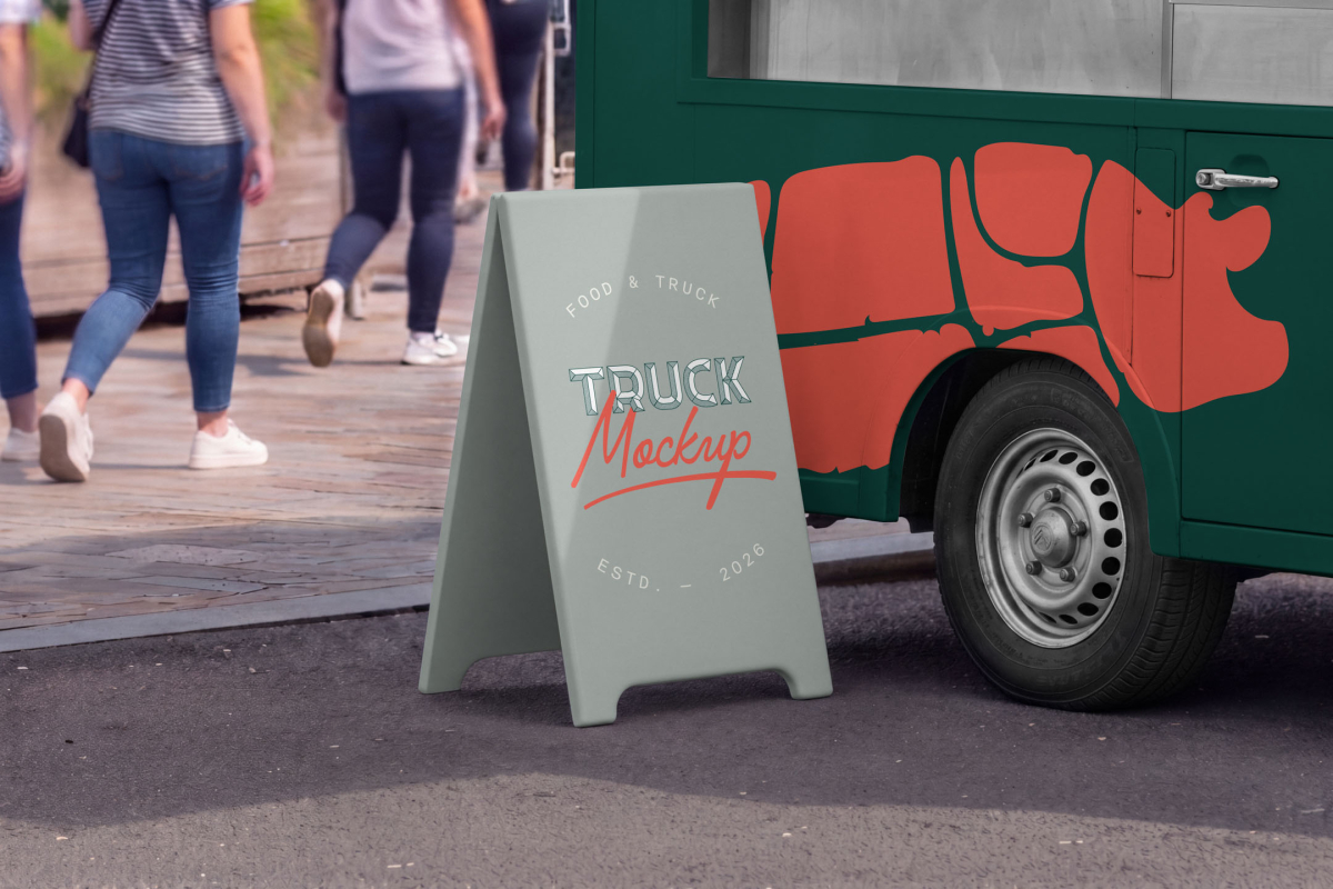 Close-up of a grey A-frame sidewalk sign standing on the pavement next to the front wheel of a dark green food truck with red graphics.