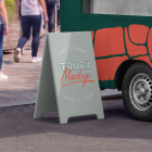Close-up of a grey A-frame sidewalk sign standing on the pavement next to the front wheel of a dark green food truck with red graphics.