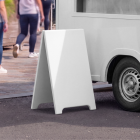 Close-up mockup of a blank white A-board sidewalk sign next to the wheel of a food truck on a paved walkway.