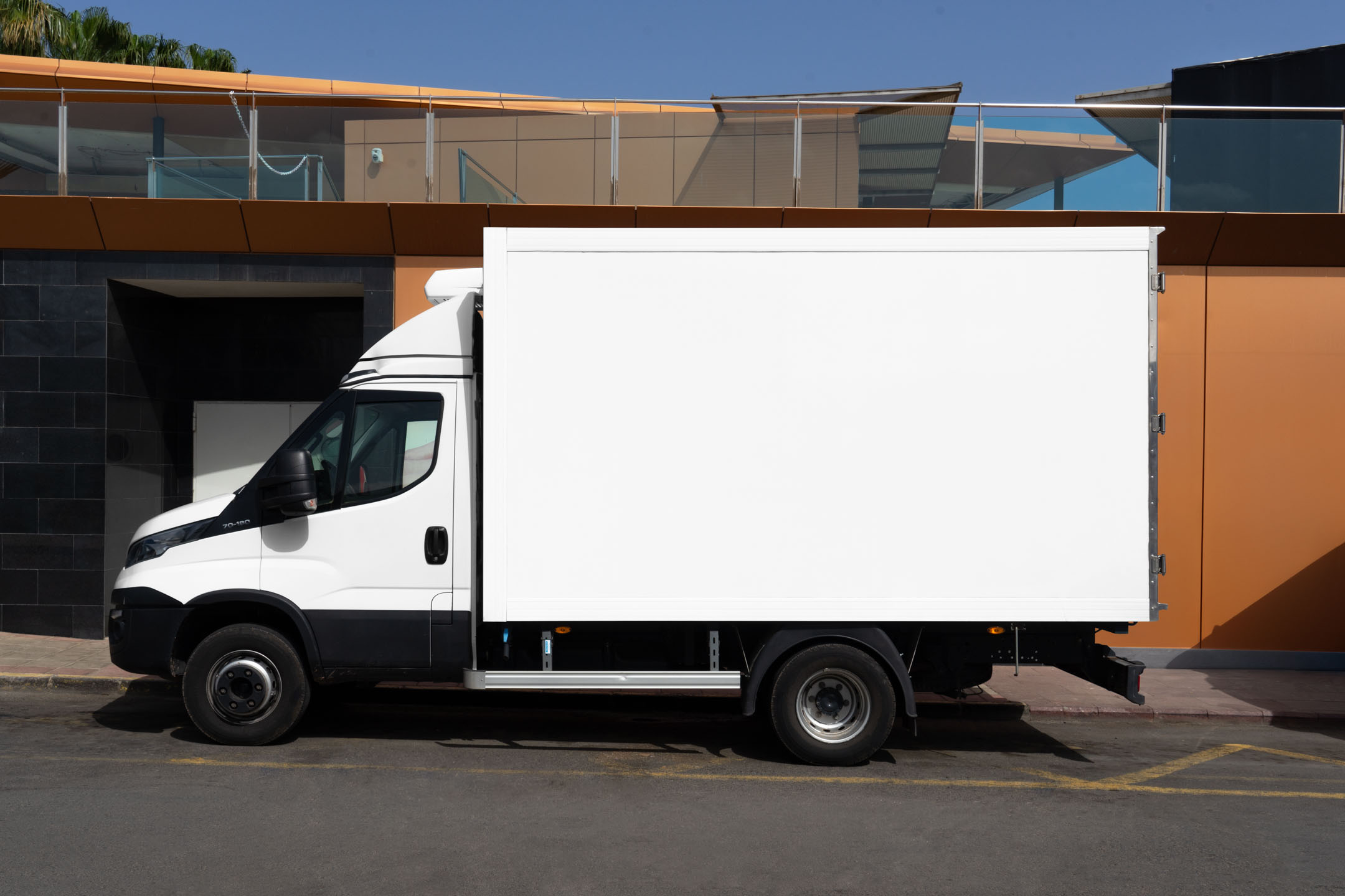 Side view profile of a white commercial cargo box truck parked against a modern building with metallic and orange panels.
