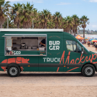 Side view of a dark green food truck mockup with an open service window, parked near a sandy beach with palm trees and people enjoying a sunny day in the background.
