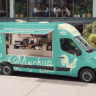 High-angle view of a turquoise food truck mockup with an open service window and a chef preparing burgers inside, parked along an urban street near a modern office building.