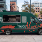 Profile view of a dark green food truck mockup with an open service window and a posted menu, parked along a city street curb in front of glass office buildings.