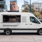 Side profile of a white Mercedes Sprinter food truck mockup with open hatch parked along a city street curb.