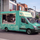 Side view of a turquoise food truck mockup with an open service window, parked on a paved city street with pedestrians walking in the background.