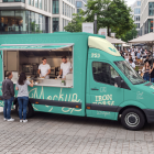 Angle view of a turquoise burger food truck mockup with an open hatch, parked on a paved pedestrian square with staff serving customers and outdoor dining tables in the background.