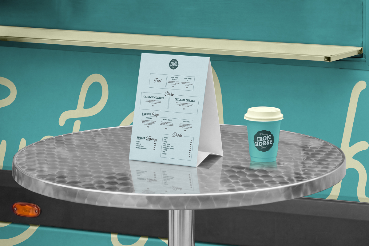 Close-up of a paper table tent menu and a matching turquoise coffee cup sitting on a round metal table in front of a turquoise food truck hatch.