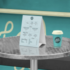 Close-up of a paper table tent menu and a matching turquoise coffee cup sitting on a round metal table in front of a turquoise food truck hatch.