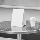 Close-up mockup of a blank white paper table tent menu and paper coffee cup on a metal table in front of a food truck.