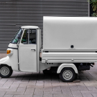Profile side view of a vintage off-white Piaggio Ape three-wheeler cargo truck parked on a paved sidewalk against a slatted metallic wall.