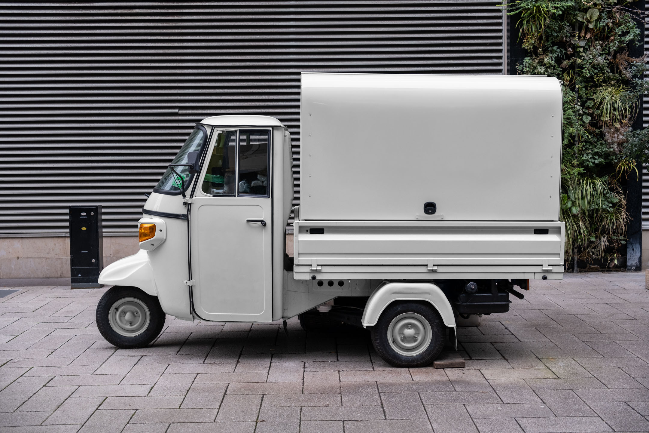 Profile side view of a vintage off-white Piaggio Ape three-wheeler cargo truck parked on a paved sidewalk against a slatted metallic wall.