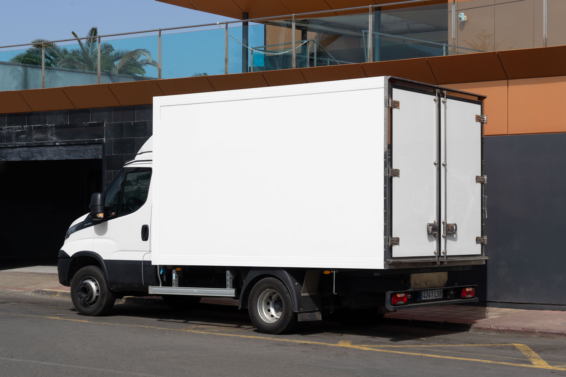 Rear three-quarter view of a white delivery box truck with double back doors parked on an asphalt street near a modern structure.