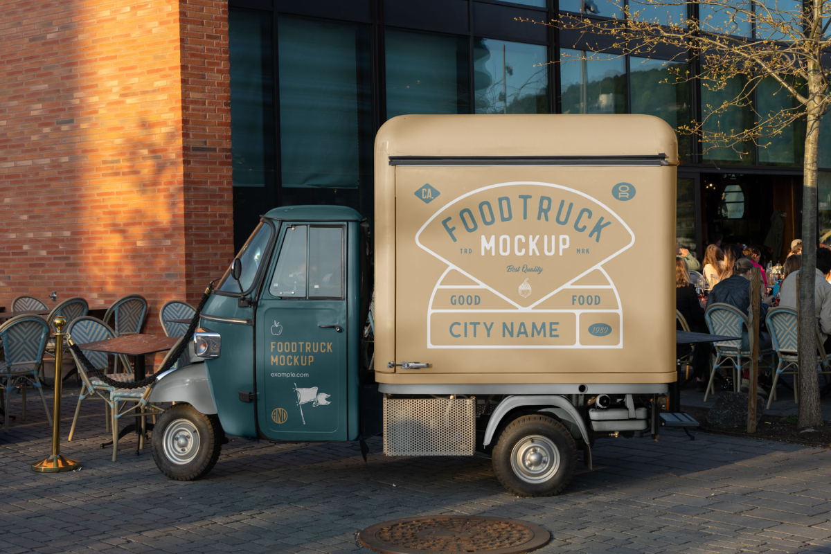 Side view of a vintage teal and beige Piaggio Ape three-wheeler food truck parked on a paved plaza near outdoor cafe seating.