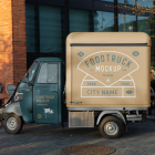 Side view of a vintage teal and beige Piaggio Ape three-wheeler food truck parked on a paved plaza near outdoor cafe seating.