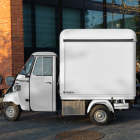 Side profile of an off-white Piaggio Ape three-wheeler food truck mockup parked on a paved plaza next to outdoor cafe seating.