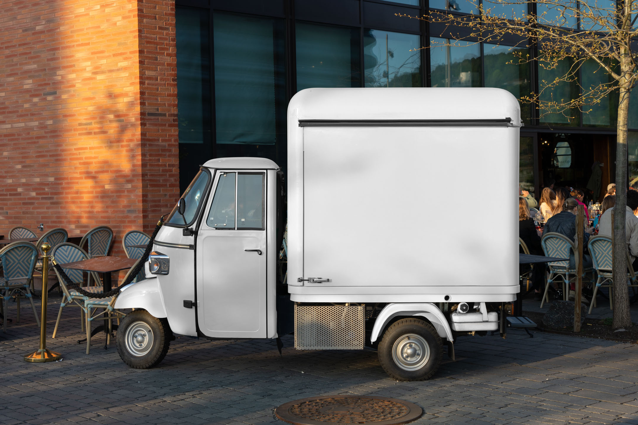 Side profile of an off-white Piaggio Ape three-wheeler food truck mockup parked on a paved plaza next to outdoor cafe seating.