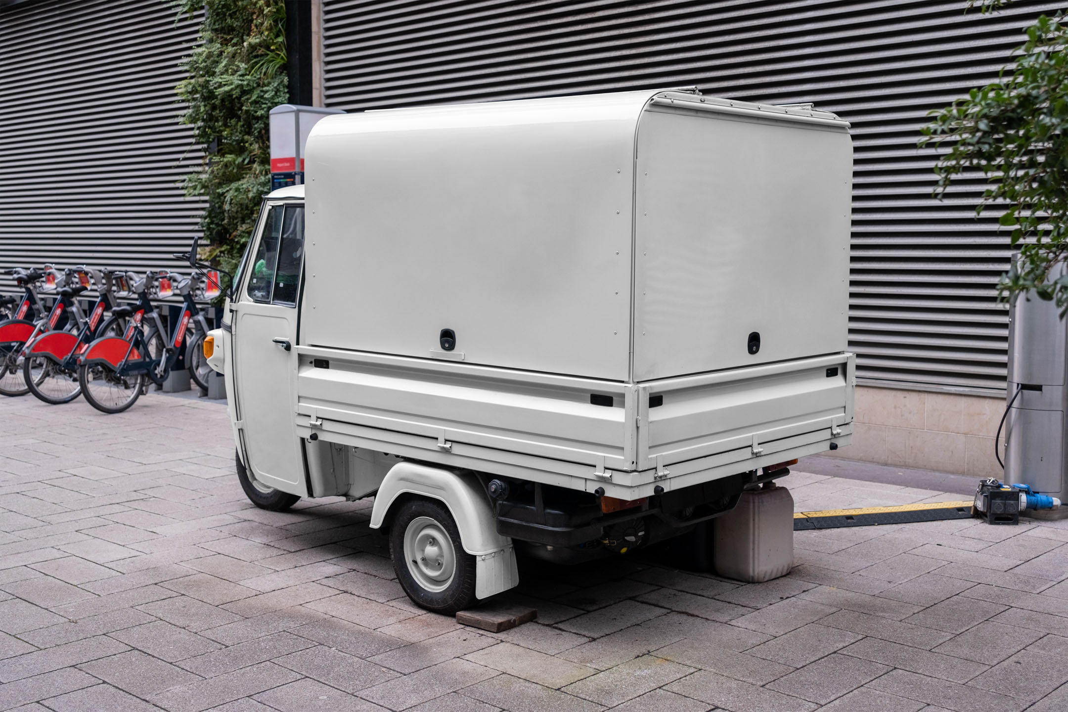Rear angle view of a vintage off-white Piaggio Ape three-wheeled cargo truck parked on a paved pedestrian walkway.