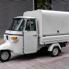 Front three-quarter angle of a small off-white Piaggio Ape three-wheeler commercial truck parked on a paved sidewalk.