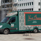 Side profile of a dark green Mercedes Sprinter food truck mockup with red framing and burger icons, parked in an open city plaza.