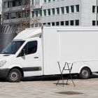 Side angle view of a white modern Mercedes Sprinter food truck mockup with high-top roof, parked in a city plaza with small foldable tables in front.