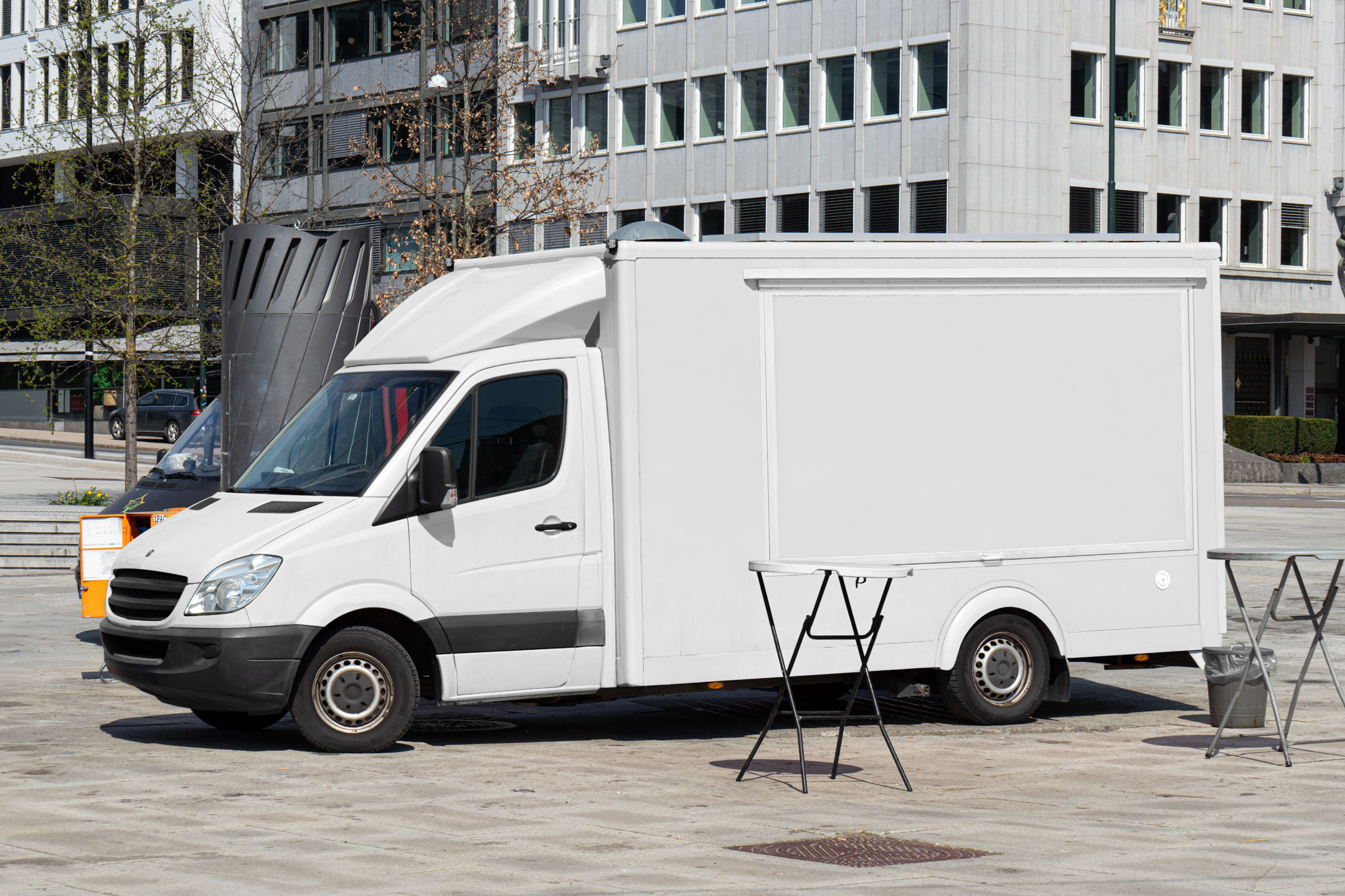 Side angle view of a white modern Mercedes Sprinter food truck mockup with high-top roof, parked in a city plaza with small foldable tables in front.