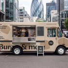 Side profile of a beige food truck mockup parked on a city street in front of modern skyscrapers, featuring an open service hatch with staff preparing food inside.