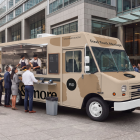Perspective view of a beige food truck mockup parked outside a corporate office building, showing a queue of business professionals ordering food from staff inside.