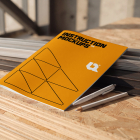 Orange instruction booklet resting on OSB boards beside two pens at a job site.
