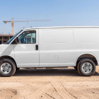 Side view of a plain white delivery cargo van parked at a dusty construction site.