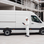 A construction worker in a white uniform holding a clipboard next to a white cargo van at a building site.