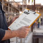 Close-up of a worker holding an orange clipboard with a document at a construction site.