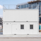 Front view of a two-story white modular site office container with a top railing at a construction site.