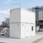 Perspective view of a two-story white site office container with external metal stairs at a construction site.