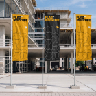 Three vertical promotional flags standing on concrete pedestals in front of a multi-story building under construction.