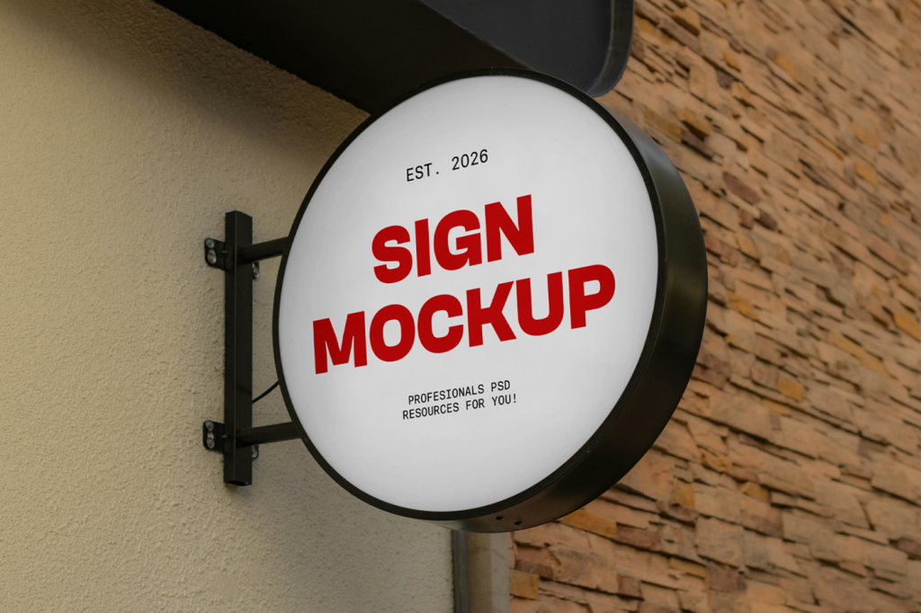 A low-angle shot of a large, circular light box sign mounted on an exterior building wall. The mockup features a blank, illuminated white face framed in matte black metal with a matching wall bracket, set against a background of light stucco and a stone tile facade. Free Big Circle Signage Mockup