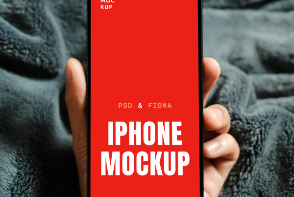 A hand holds a modern smartphone with a blank white screen, likely an iPhone with a dynamic island notch, against a backdrop of a soft, textured dark grey blanket. Free Hand Holding by iPhone PSD Mockup