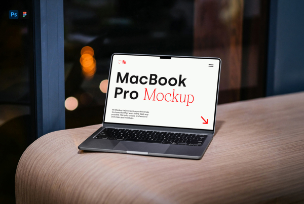 A dark grey laptop with a blank white screen resting on a light wooden surface next to a window with warm ambient background lights. Free MacBook Pro Laying on Wood Mockup