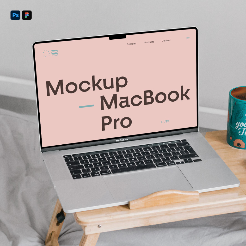 A silver MacBook laptop with a blank white screen is open on a small, folding wooden bed desk. Next to it on the desk are a teal coffee mug with the text "See you next Tuesday" and a white computer mouse. The setup is placed on grey bedding in a bedroom setting. Free MacBook Pro in Bed Mockup