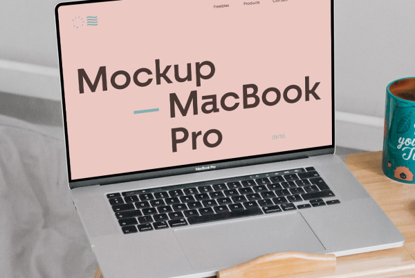 A silver MacBook laptop with a blank white screen is open on a small, folding wooden bed desk. Next to it on the desk are a teal coffee mug with the text "See you next Tuesday" and a white computer mouse. The setup is placed on grey bedding in a bedroom setting. Free MacBook Pro in Bed Mockup