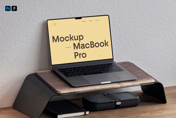 A modern desk setup mockup featuring a dark grey MacBook Pro with an open, blank white screen resting on a wooden and metal monitor riser. Beneath the stand sits a black notebook and an accessory case, set against a light-textured white wall in natural indoor lighting. Free MacBook Pro on Wooden Pad Mockup