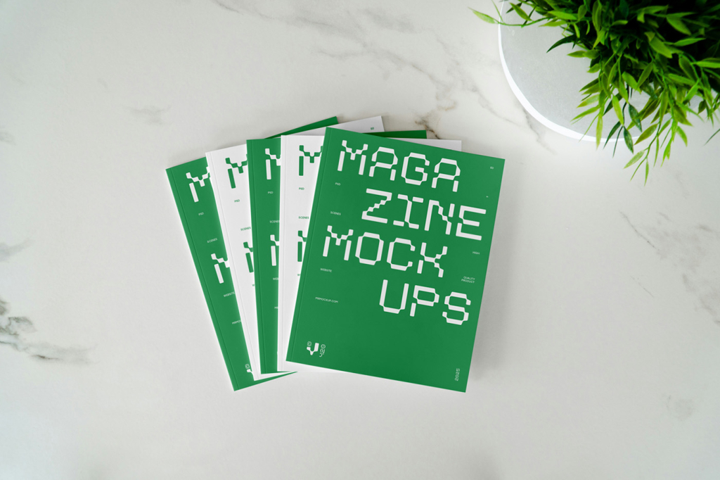 op-down view of several blank white magazines or brochures neatly fanned out on a white marble surface, with a small green potted plant in the upper right corner. Free Magazine Cover Laying on Marble Mockups