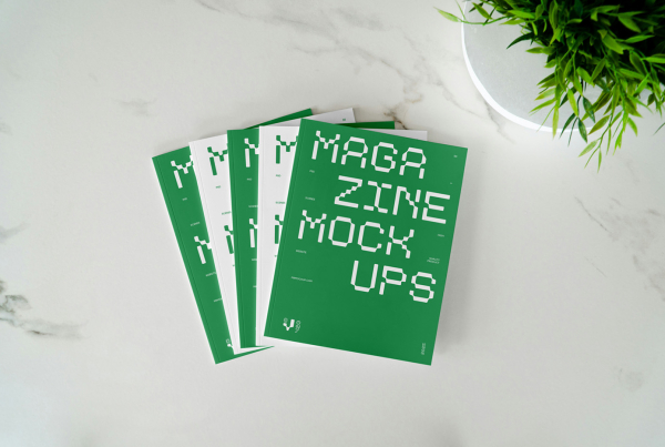 op-down view of several blank white magazines or brochures neatly fanned out on a white marble surface, with a small green potted plant in the upper right corner. Free Magazine Cover Laying on Marble Mockups