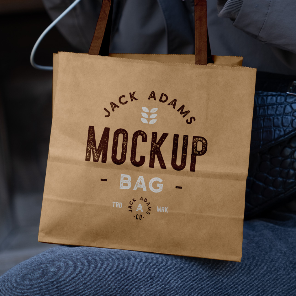 A person holding a plain, light brown mini paper shopping bag by its handles, resting it on their lap while wearing dark jeans and a grey jacket. Free Mini Paper Shopping Bag Mockup