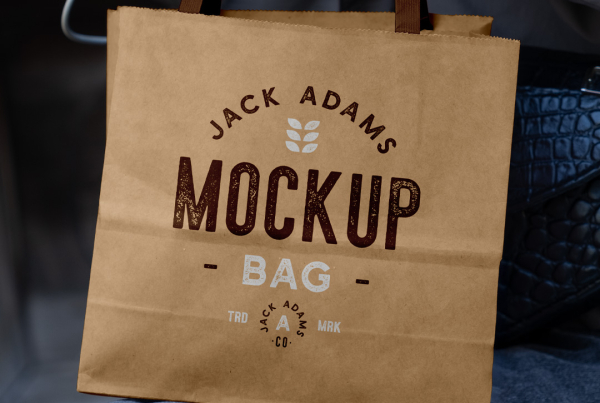 A person holding a plain, light brown mini paper shopping bag by its handles, resting it on their lap while wearing dark jeans and a grey jacket. Free Mini Paper Shopping Bag Mockup
