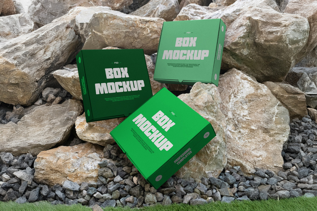 Three blank square cardboard boxes arranged dynamically and propped against a large pile of rugged natural rocks and stones. Free Three Boxes Mockups